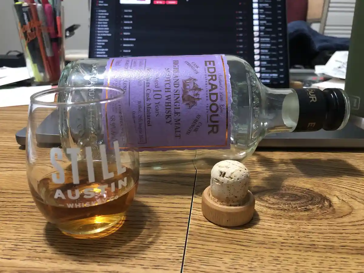 A glass of amber-colored whiskey labeled 'STILL AUSTIN WHISKEY CO' sits on a wooden desk next to an empty Edradour Highland…