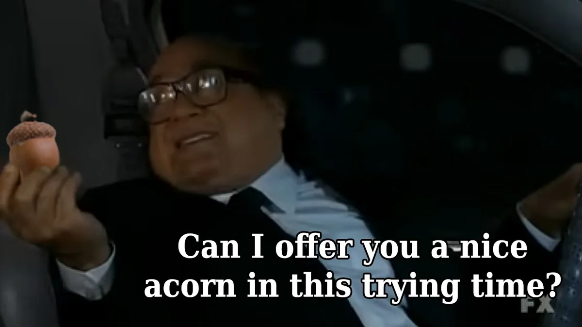 A man in a suit and glasses sits in a car, holding up an acorn while looking at the camera. White text overlaid on the image…