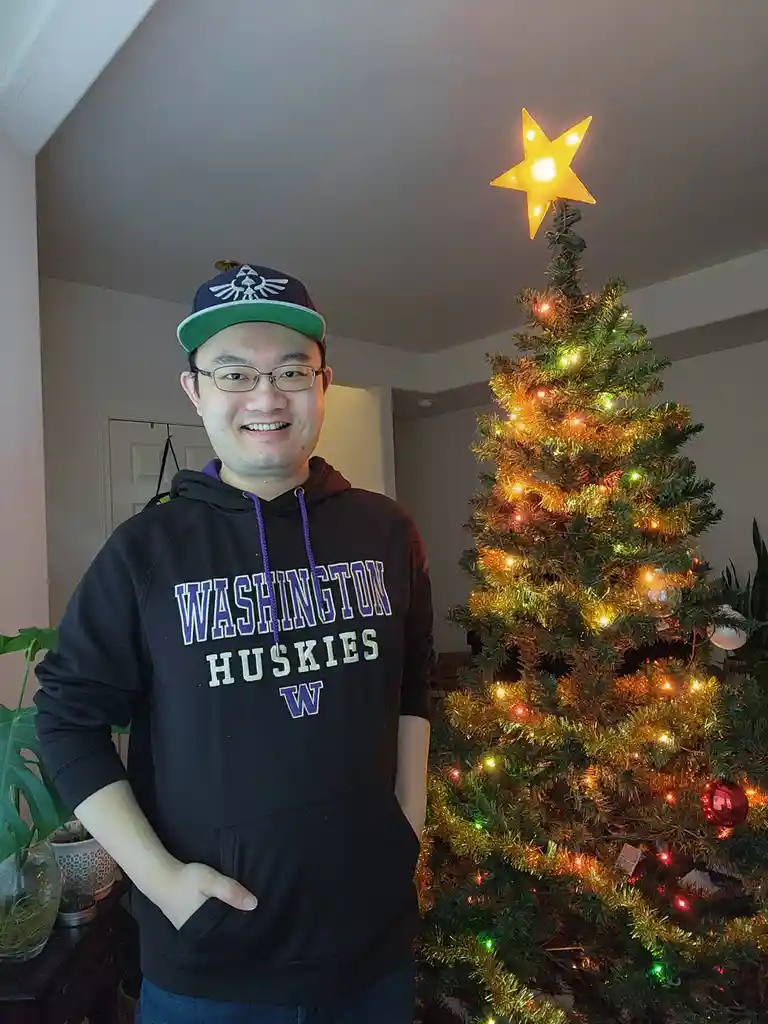 A smiling man wearing a Washington Huskies hoodie and a Zelda-themed cap stands beside a decorated Christmas tree with…