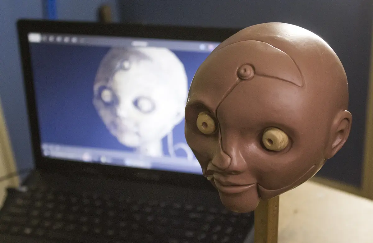 A brown, featureless robot head with large, yellowish eyes and a subtle smile sits on a stand in front of a laptop. The…