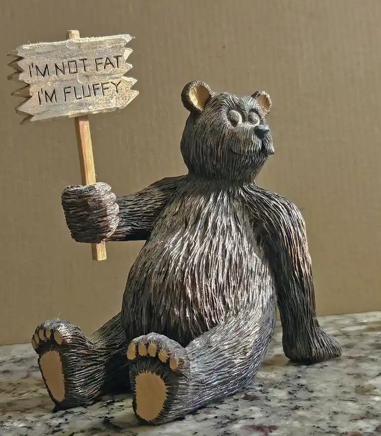 A carved wooden bear figurine sits on a marble-like surface holding a wooden sign that reads 'I'M NOT FAT I'M FLUFFY'. The…