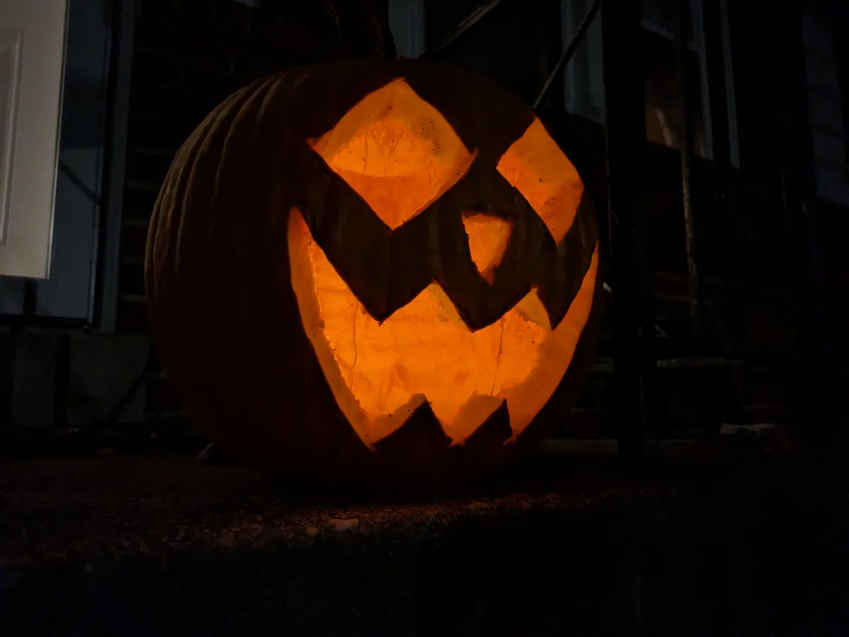 A large, carved pumpkin with a glowing orange interior sits on a porch at night, its face illuminated with triangular eyes…