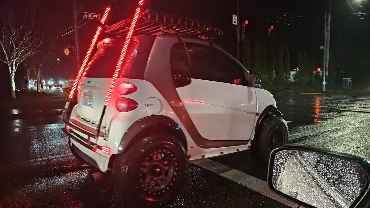 A white Smart car is stopped at a rainy night intersection, modified with a roof rack and bright red LED lights forming a…
