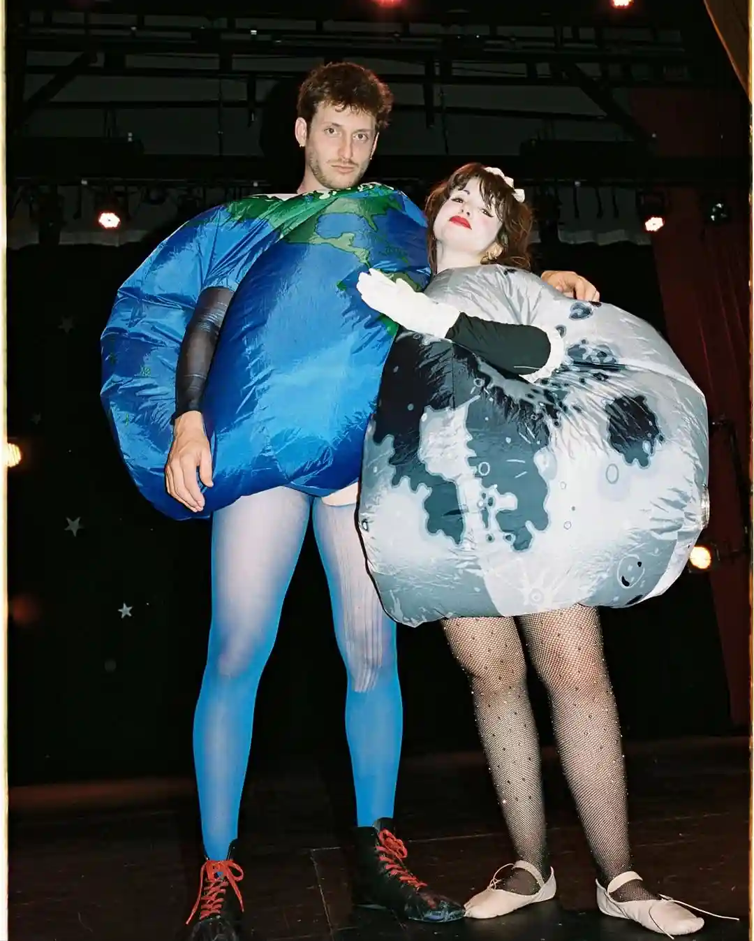 A man and a woman pose on a stage, both wearing large inflatable costumes: the man as Earth with blue oceans and green…