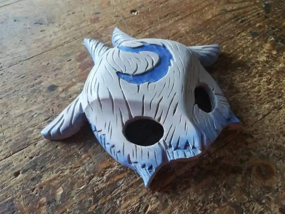 A handmade, light blue clay mask with carved horns, leaf-like patterns, and two eye holes rests on a weathered wooden…
