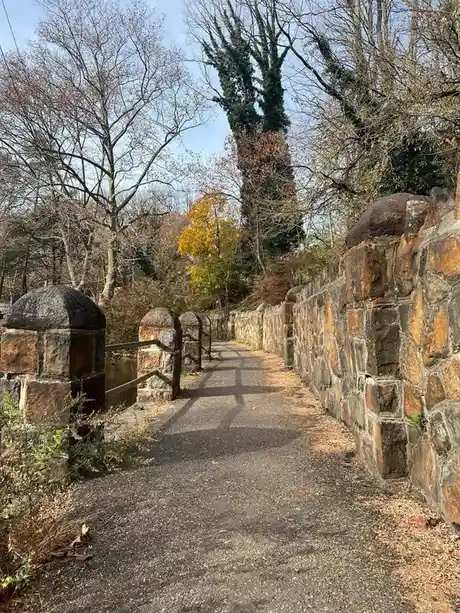 A paved path winds through a park flanked by a stone wall on the right and a low stone fence on the left, leading toward a…