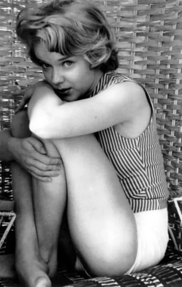 A black-and-white photo of a young girl with short, wavy hair, wearing a striped tank top and shorts, sitting on a wicker…