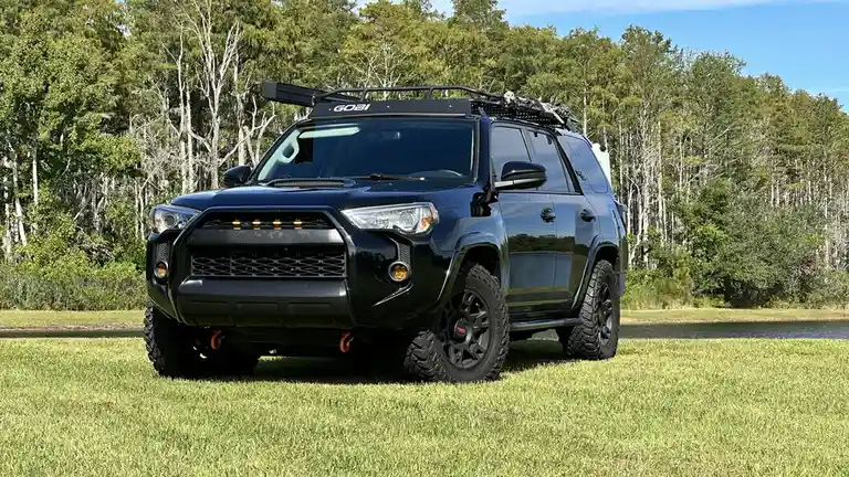 A black, heavily modified Toyota 4Runner is parked on a grassy field with a forested background. The vehicle features a roof…