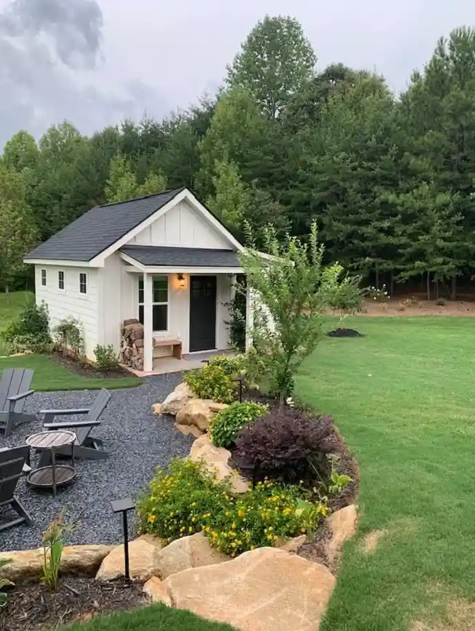 A small, white, A-frame style house with a black roof sits in a lush green yard surrounded by trees. A gravel patio with…