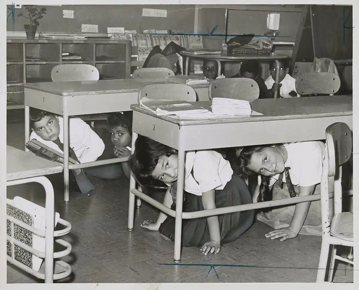 A black-and-white photo shows several children hiding under their desks in a classroom, peeking out with wide eyes. Some are…