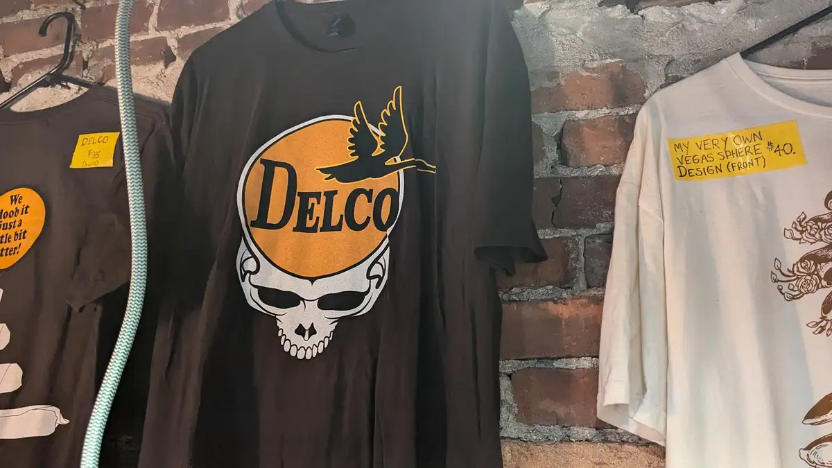 Three t-shirts hang on a brick wall, one prominently features a skull with 'DELCO' and a bird, another has a yellow tag…