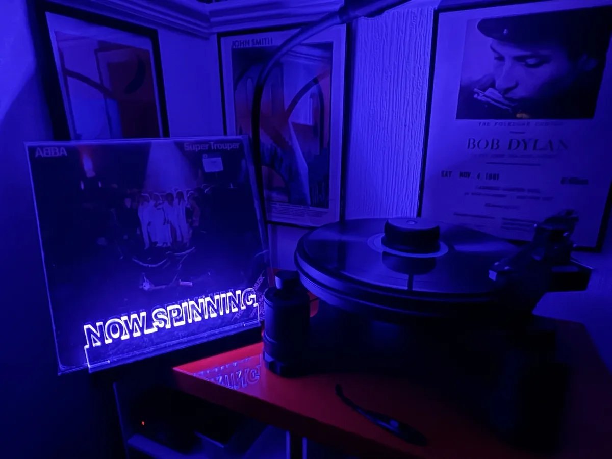 A record player sits under blue light with an ABBA 'Super Trouper' album cover beside it, displaying glowing text that reads…