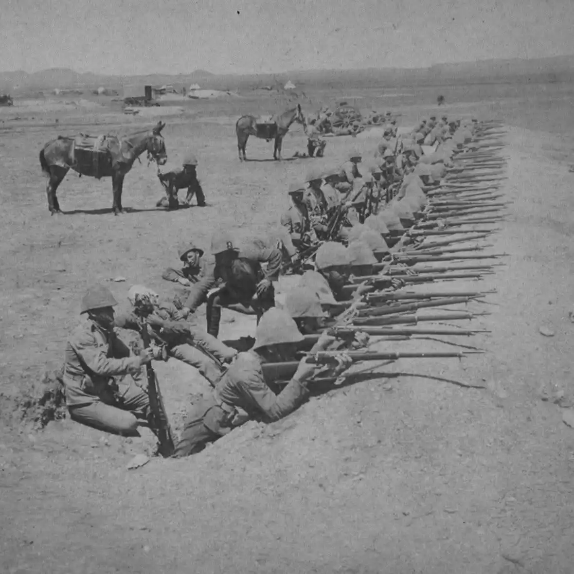 A black-and-white historical photo shows soldiers in helmets lying prone in shallow trenches, aiming rifles in a long…