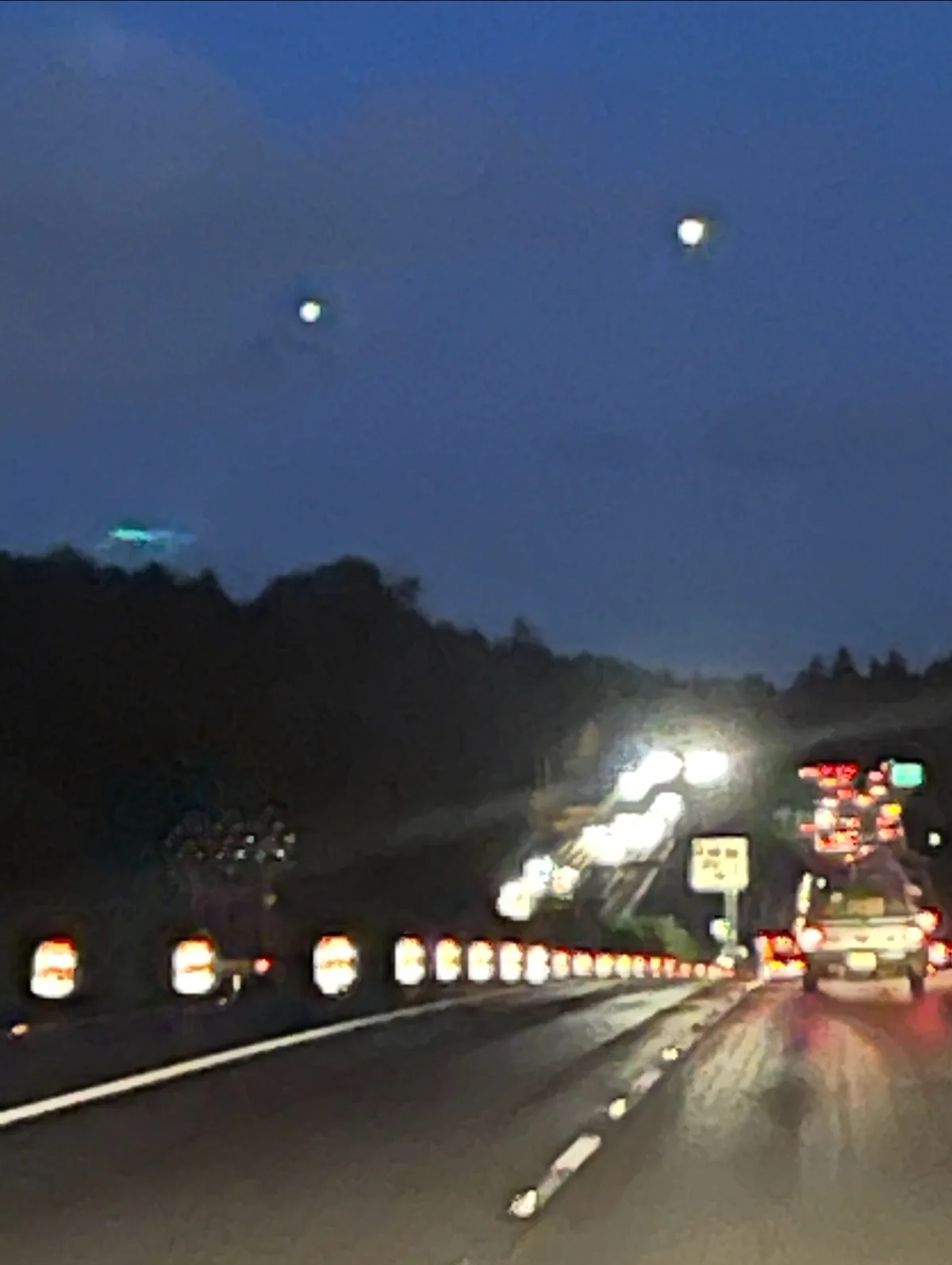 A blurry nighttime highway photo shows two bright, circular lights in the dark sky above the road, with car taillights and…