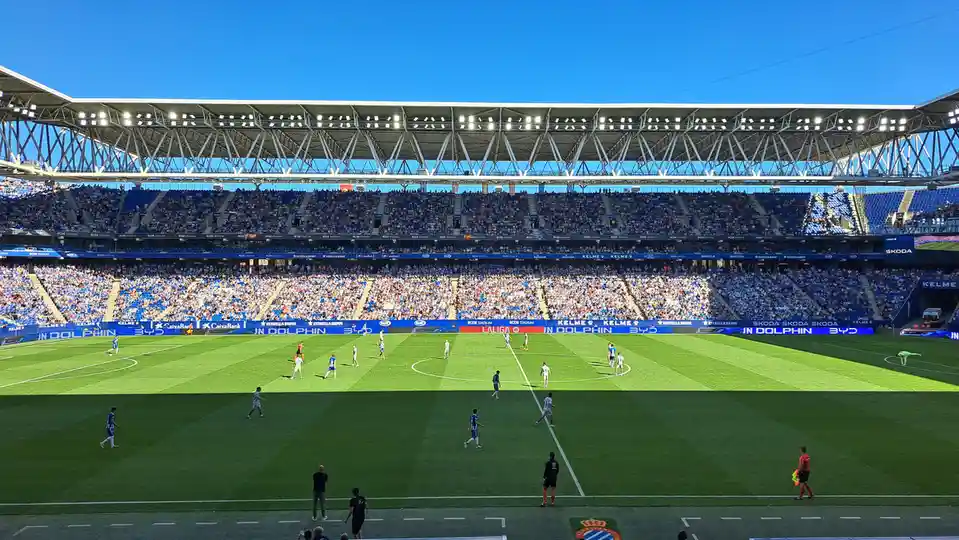 A wide shot of a packed soccer stadium under a clear blue sky, with players on the green field and fans in the stands…