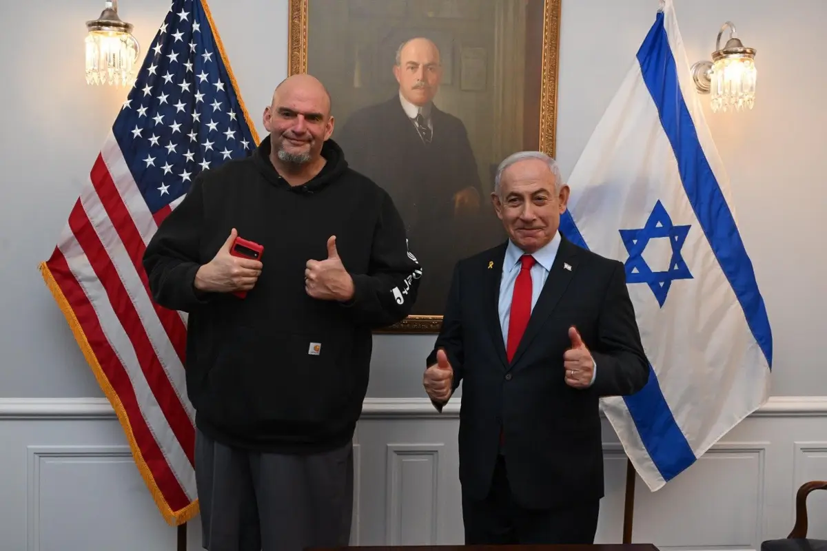 Two men stand side-by-side in a formal room with an American flag on the left and an Israeli flag on the right, both giving…