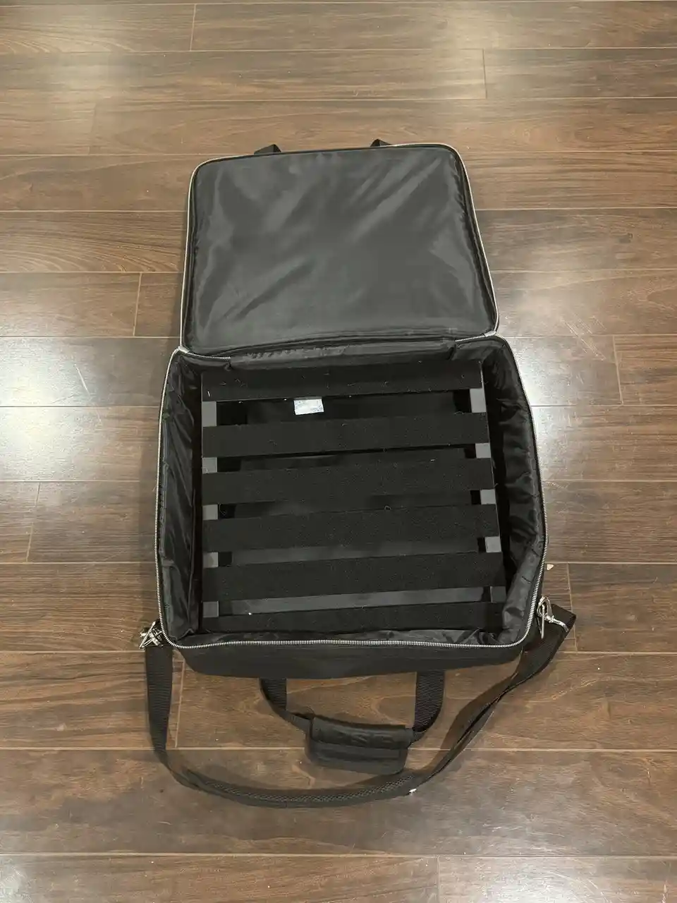 An overhead shot shows an open black soft-sided carrying case resting on a wooden floor, revealing its empty interior lined…