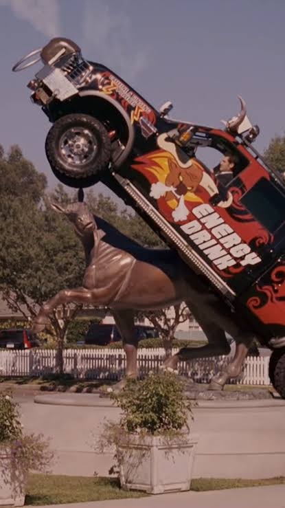 A large, red and black energy drink branded truck is balanced precariously on the back of a bronze bull statue, with the…