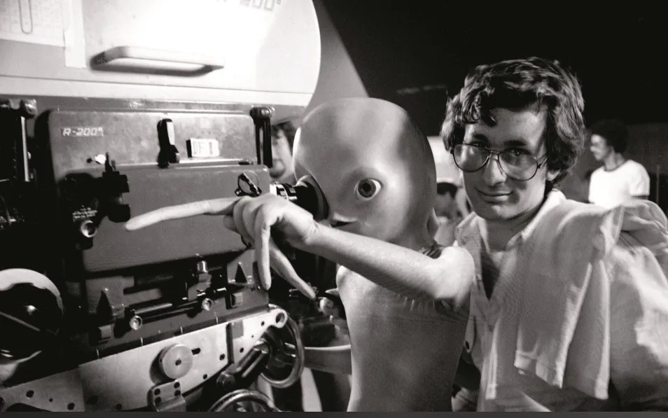 A black-and-white photo shows a man with glasses and a white shirt standing beside a large film camera labeled 'R-2001'. He…