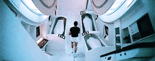 A person in a black shirt and white shorts walks down a futuristic, white, curved hallway lined with identical, empty…