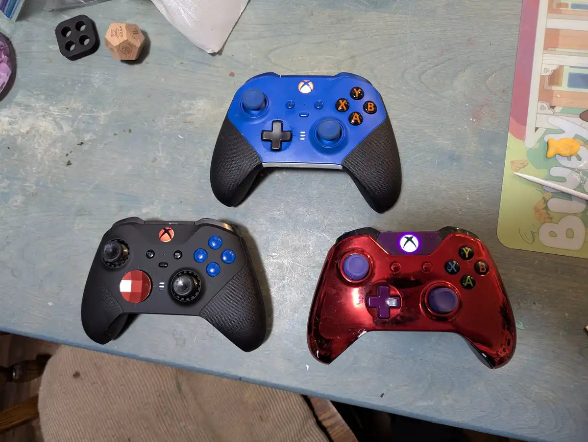 Three Xbox controllers are arranged on a worn blue table: a blue one at the top, a black one with custom red and blue…