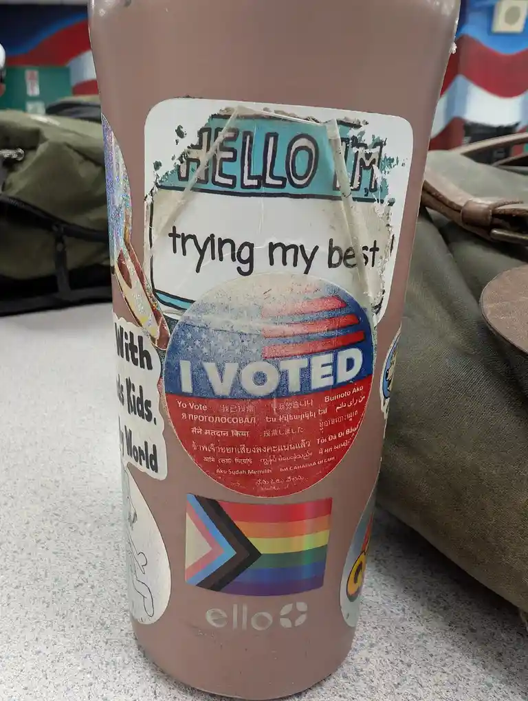 A brown reusable water bottle covered in various stickers, including a prominent 'I VOTED' sticker with multilingual text, a…