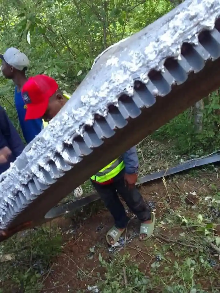 A person in a red cap and yellow safety vest stands beside a large, rusted, toothed metal rail segment lying on muddy ground…