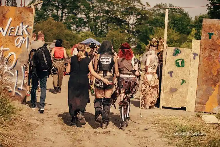 A group of people in eclectic, festival-style clothing walk away from the camera down a dirt path flanked by rustic…