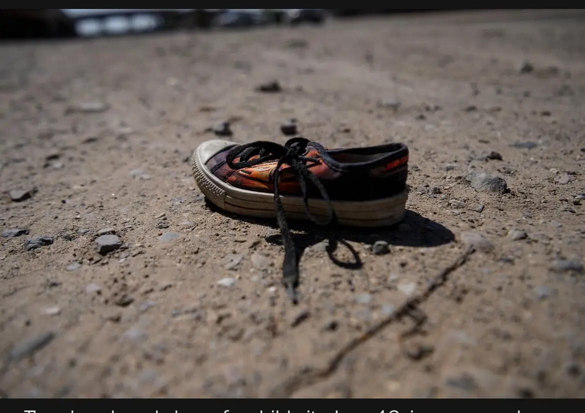 A single, worn-out child's sneaker lies abandoned on a gritty, dusty ground with small stones and gravel. The shoe…