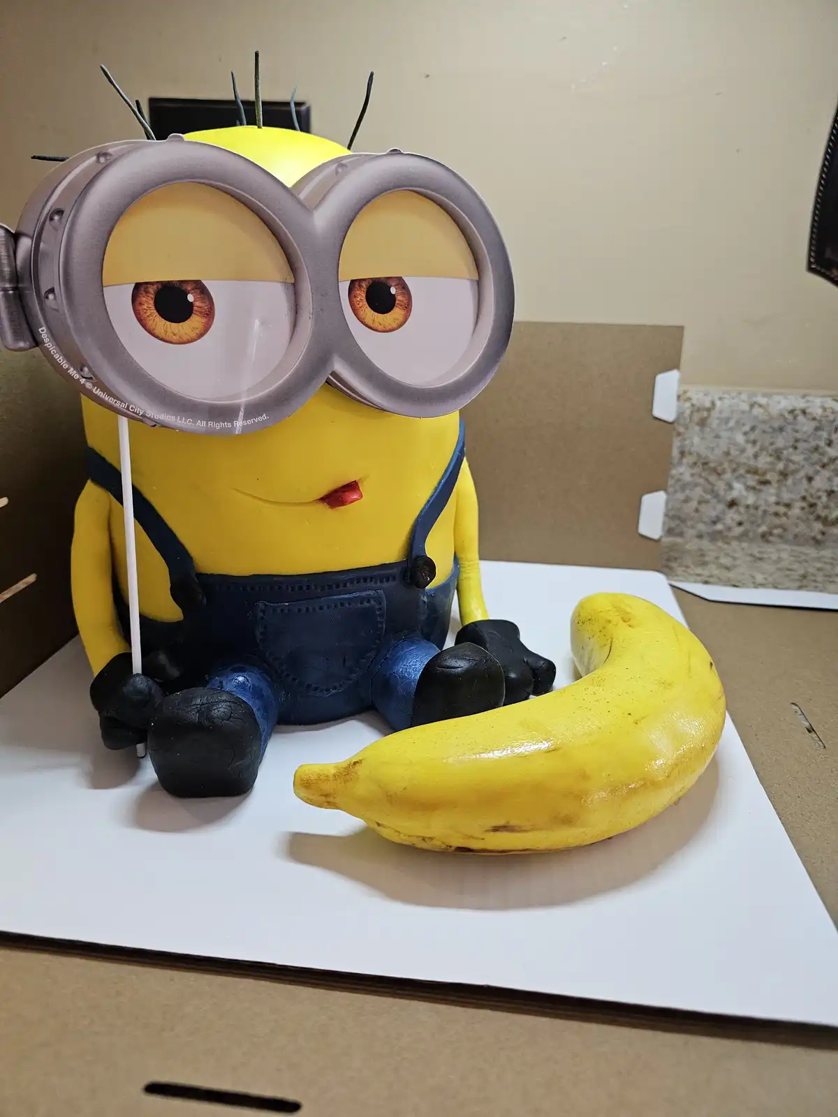 A cake shaped like a yellow Minion from Despicable Me 4, wearing its signature goggles and blue overalls, sits on a white…
