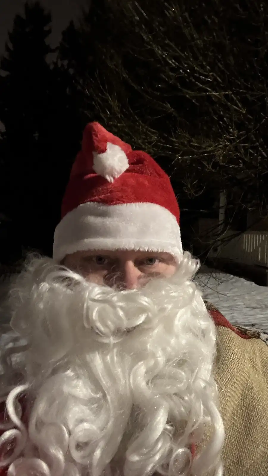 A person wearing a red and white Santa hat and a large, curly white fake beard, with only their eyes visible above the…