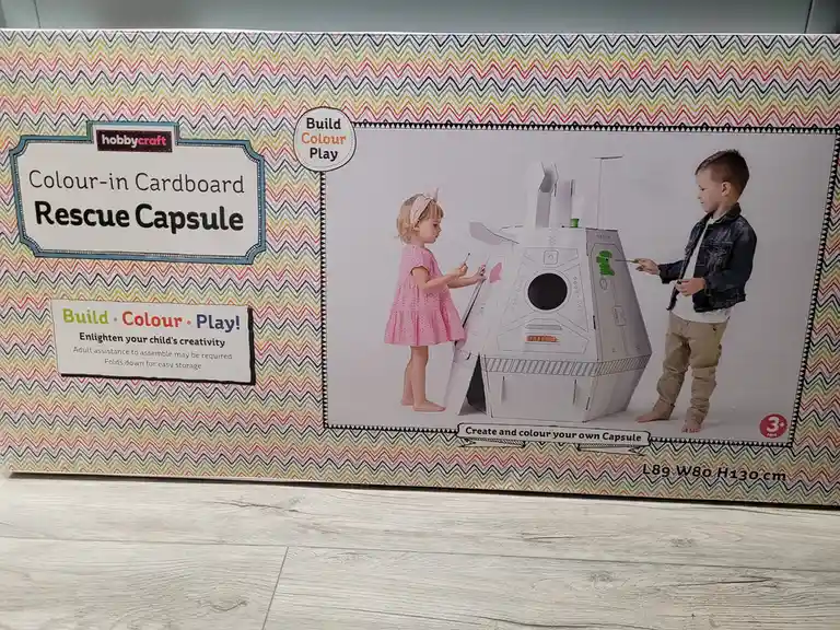 A product box for a 'hobbycraft Colour-in Cardboard Rescue Capsule' shows two children, a girl in a pink dress and a boy in…
