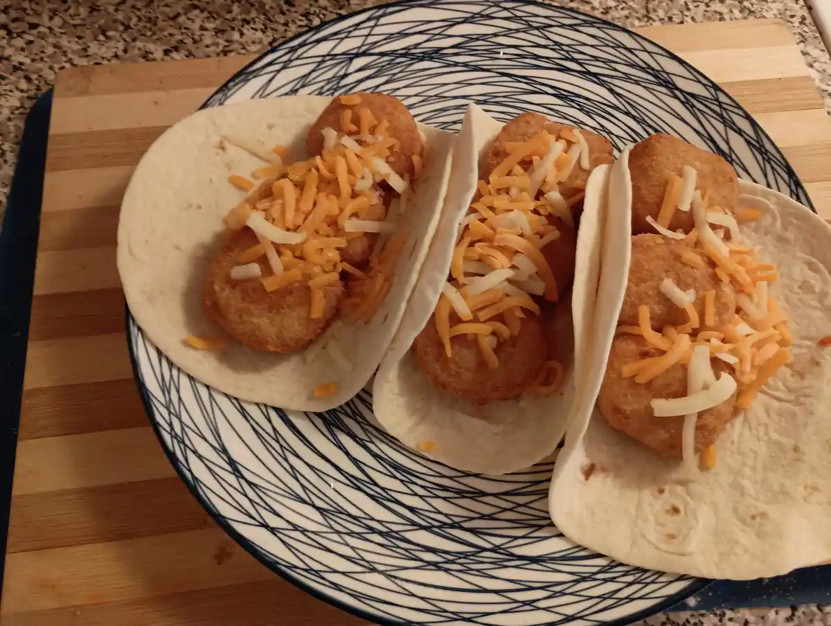 Three tacos on a blue and white patterned plate, each filled with two fried chicken nuggets and topped with shredded orange…