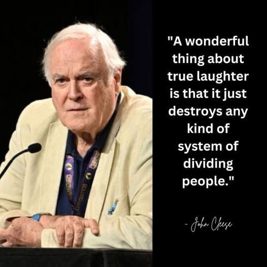 An older white man with white hair, wearing a beige blazer over a dark shirt, sits at a podium with a microphone. He has a…