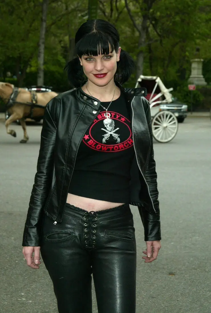 A woman with black hair and bangs, wearing a black leather jacket over a black crop top with a red skull and crossbones logo…