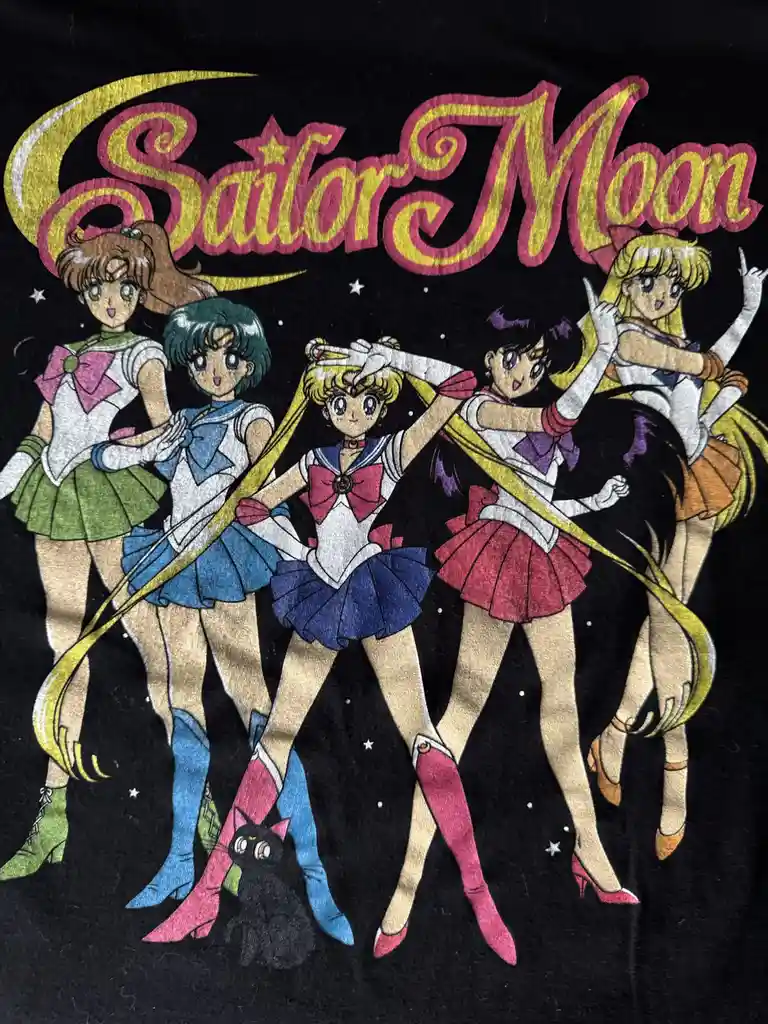 A black t-shirt features a vintage-style embroidered graphic of the five main Sailor Moon characters in their iconic sailor…