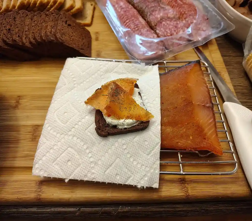 A close-up photo shows a small sandwich made with dark rye bread, cream cheese, and a thin slice of smoked salmon, garnished…