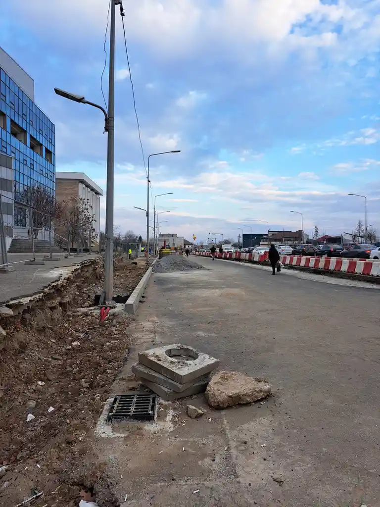 A street under construction shows a dug-up sidewalk with exposed dirt, scattered debris, and a broken concrete manhole…