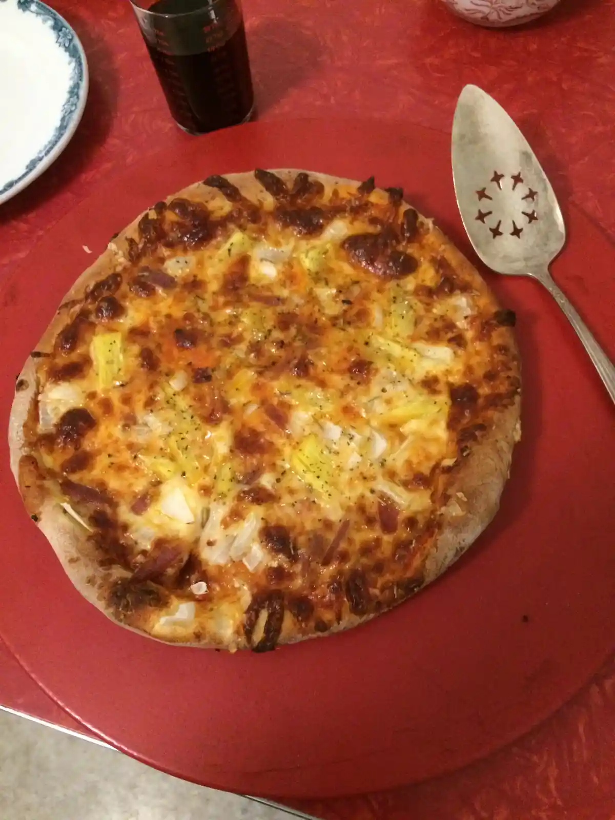 A round pizza with melted cheese, onions, and bacon sits on a red plate, next to a metal serving spoon with star-shaped…