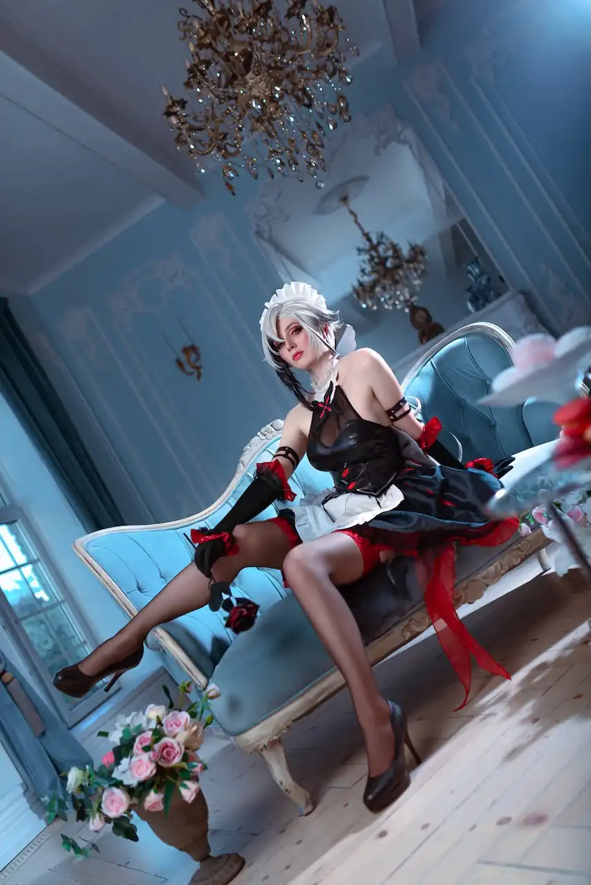 A cosplayer with silver hair and a maid outfit sits provocatively on a blue velvet chaise lounge in an ornate, light blue…