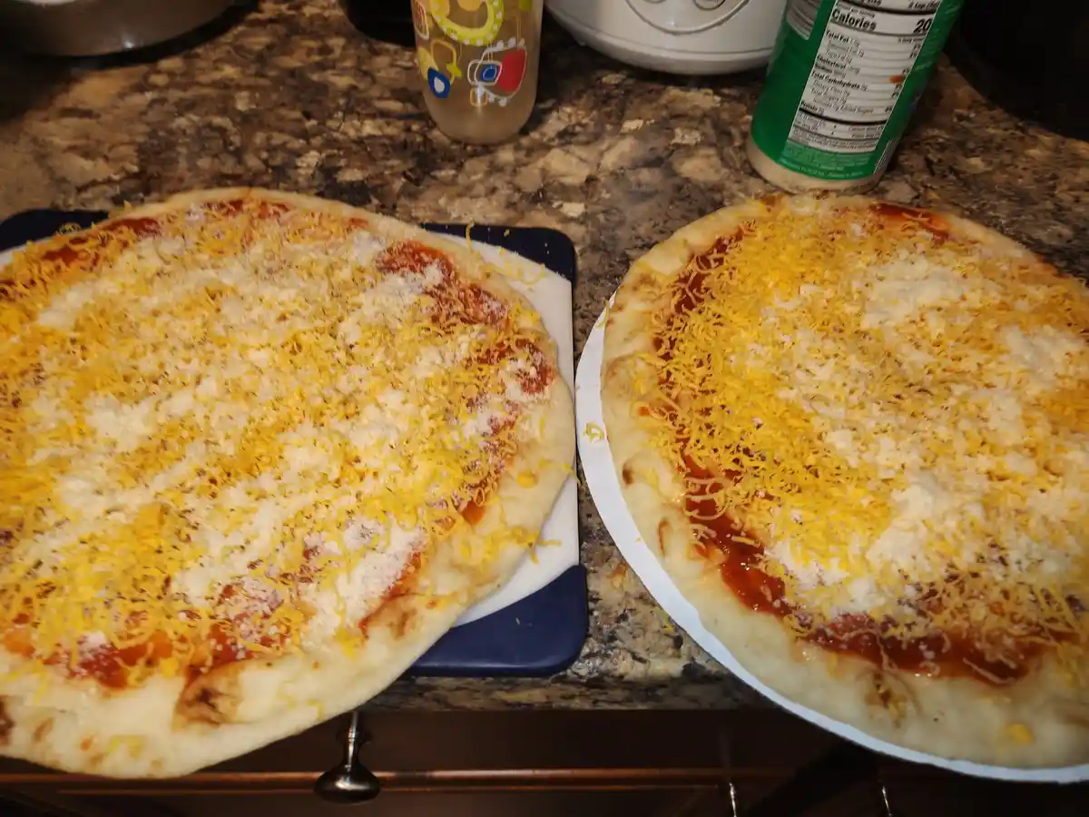 Two uncooked pizzas sit side by side on kitchen counters, both topped with tomato sauce, shredded yellow cheese, and white…