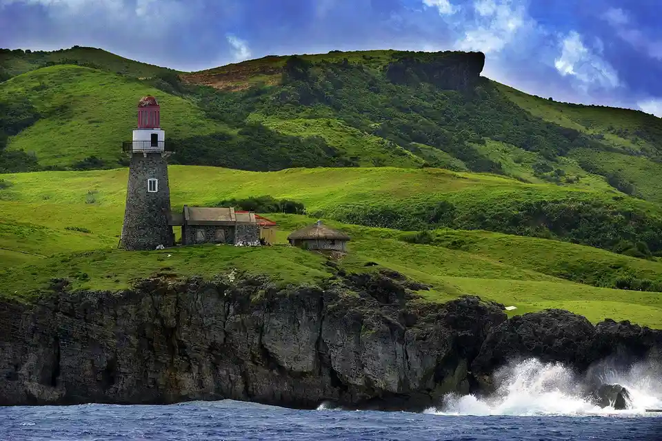 A scenic photo of a stone lighthouse with a red lantern room, situated on a grassy cliff overlooking crashing ocean waves…