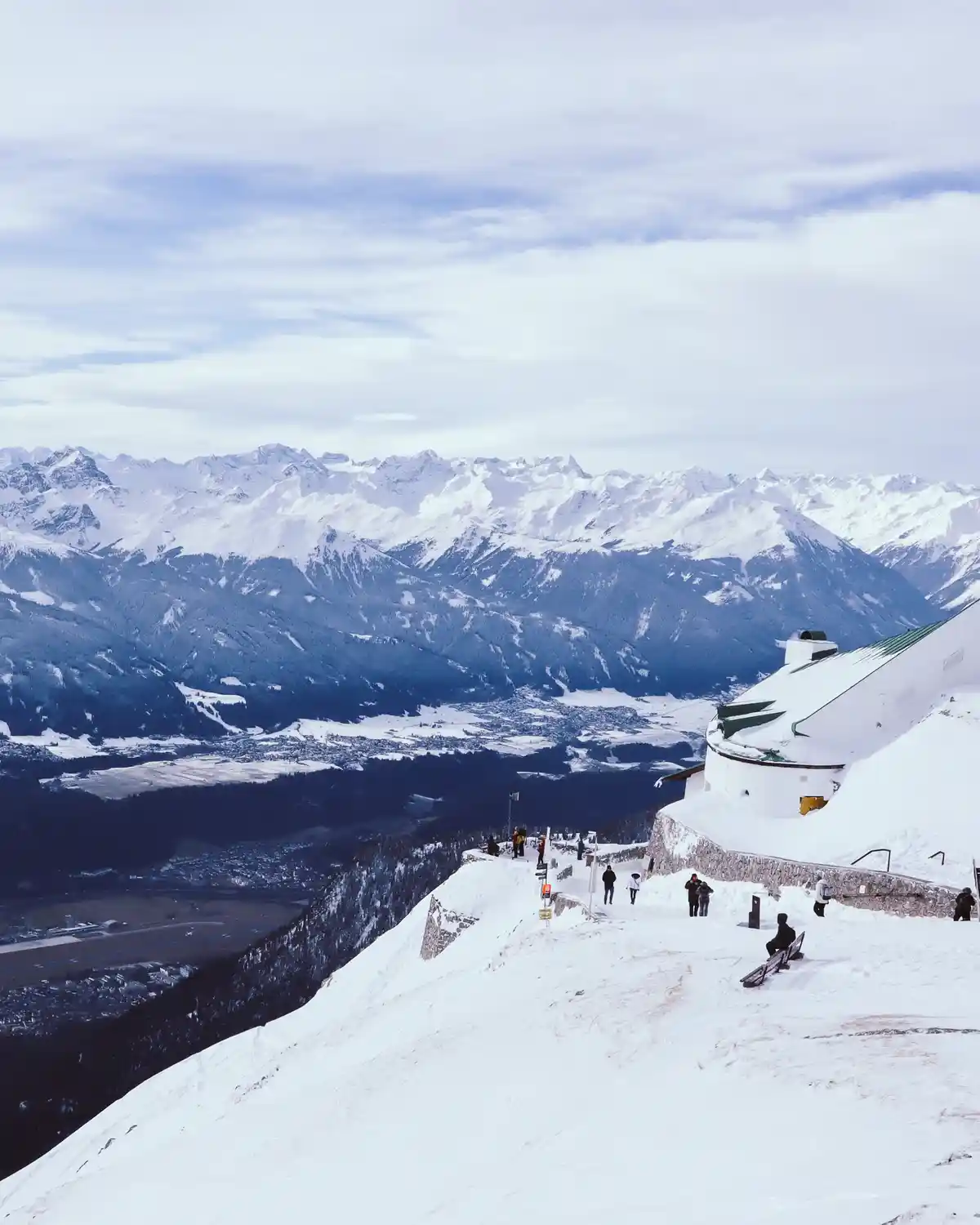 A scenic snowy mountain overlook with a white building and several people scattered around, some standing and one sitting on…