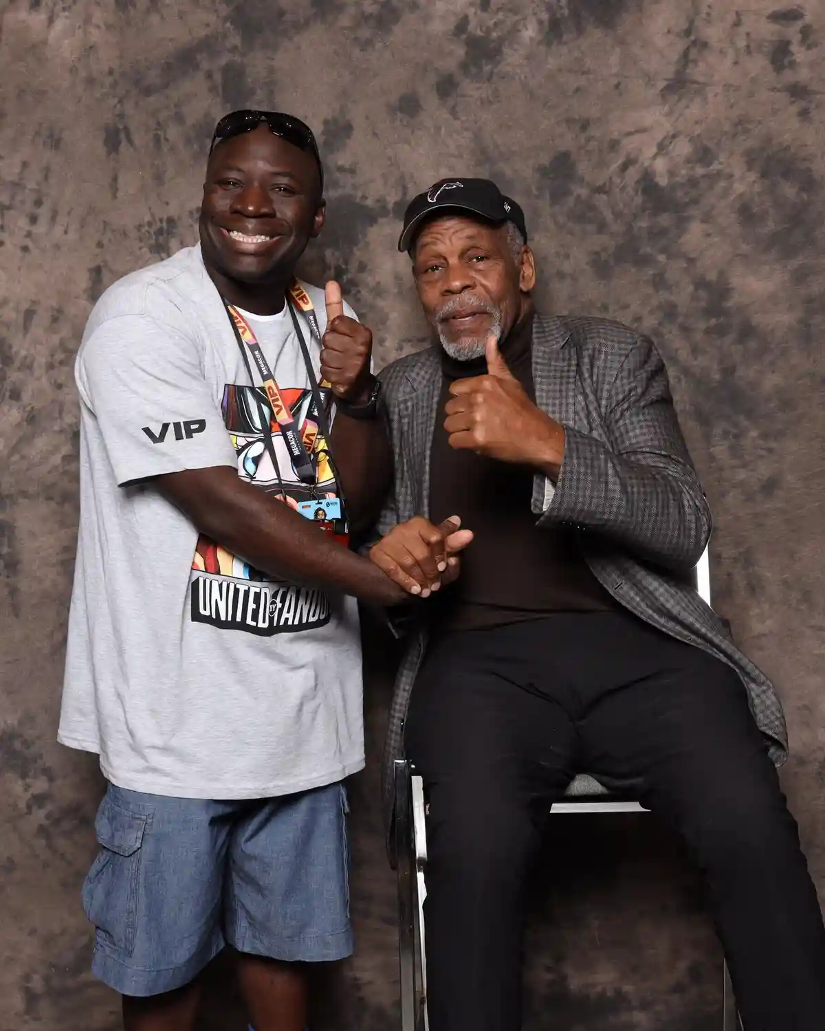 Two men pose for a photo against a mottled brown backdrop; the standing man wears a grey 'VIP' t-shirt with 'UNITED BY…
