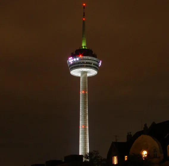 A tall, illuminated observation tower at night, lit with white, green, and red lights, stands against a dark sky with a…