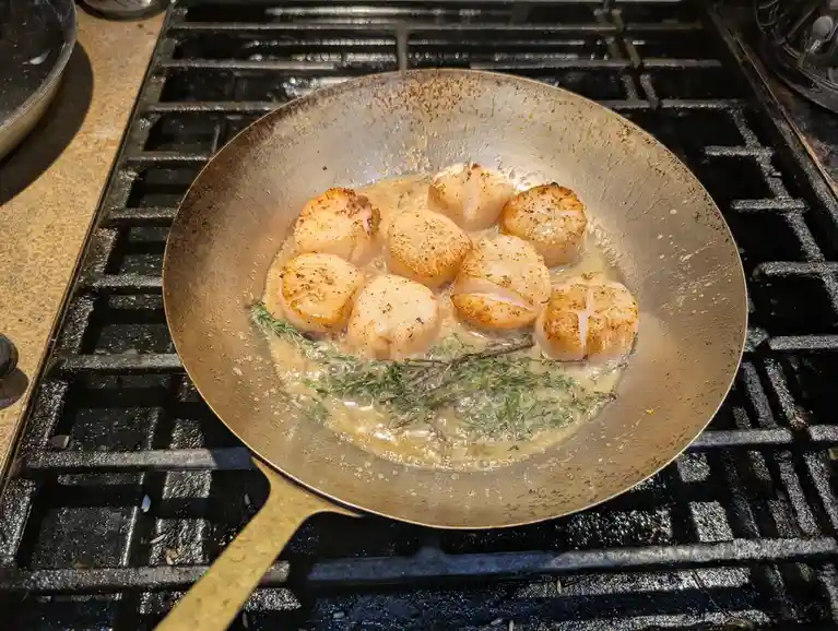 A close-up photo shows eight seared scallops sizzling in a bubbling butter and herb sauce within a well-used metal skillet…