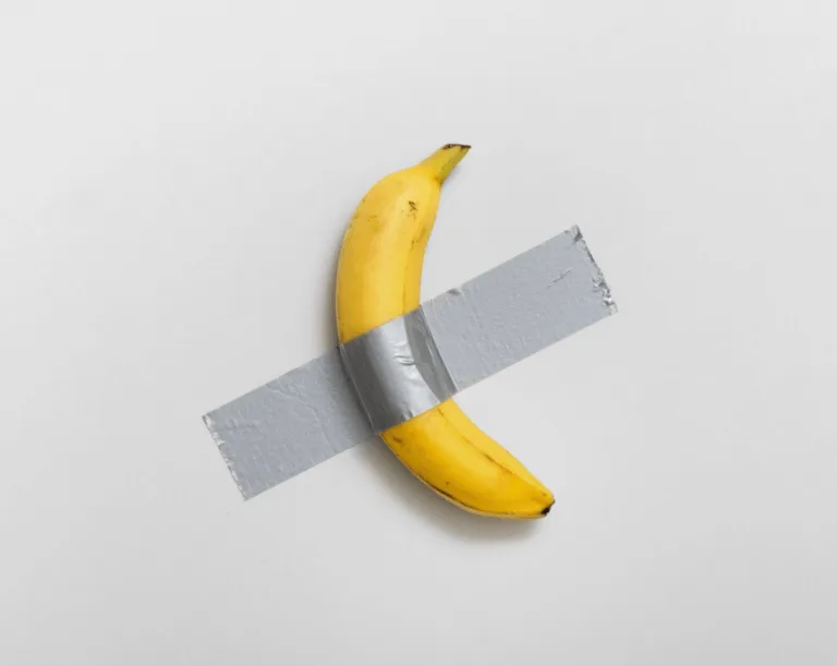 A single yellow banana lies on a plain white surface, crossed by two strips of gray duct tape forming an 'X' over its…