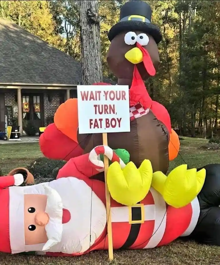 An inflatable Thanksgiving turkey wearing a pilgrim hat and holding a sign that reads 'WAIT YOUR TURN, FAT BOY' stands over…
