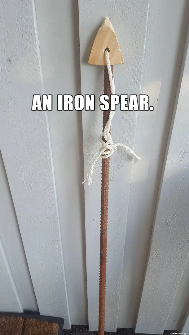 A rusty metal rod, resembling a spear shaft, is hung vertically on a white wooden door with a simple wooden arrowhead…