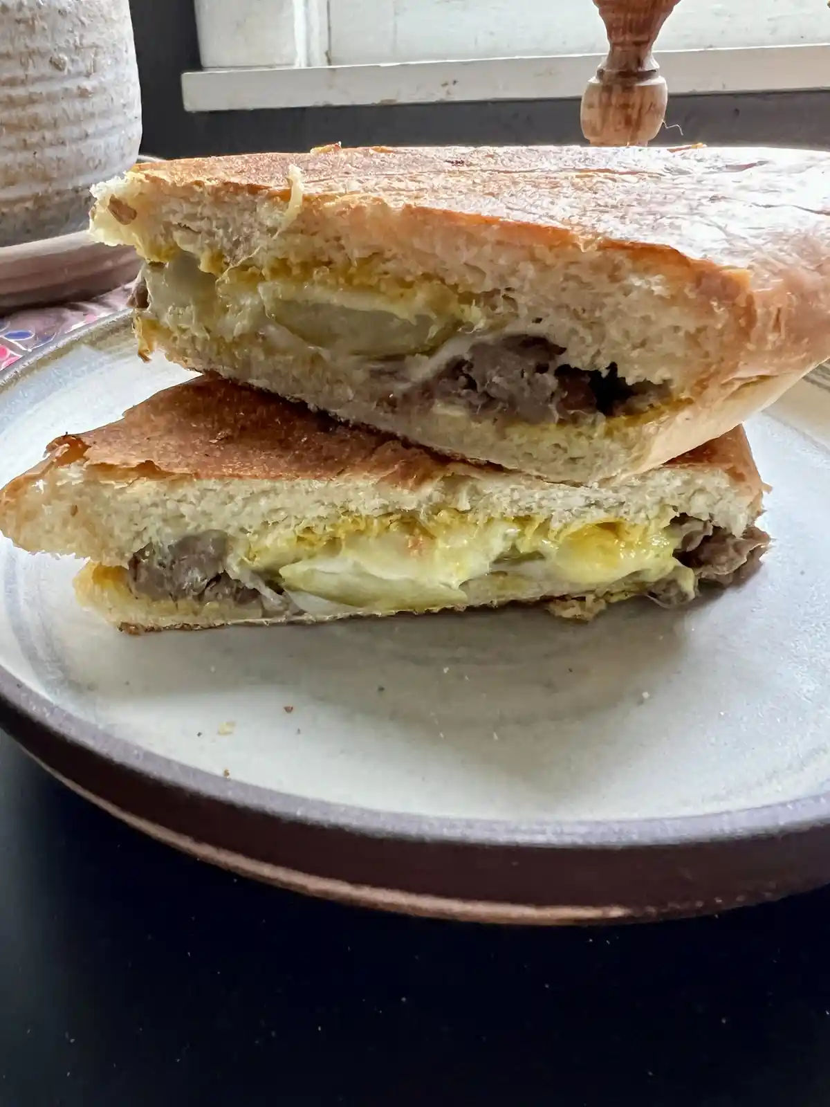 A close-up photo shows a sandwich cut in half and stacked on a ceramic plate, revealing melted cheese, scrambled egg, and…
